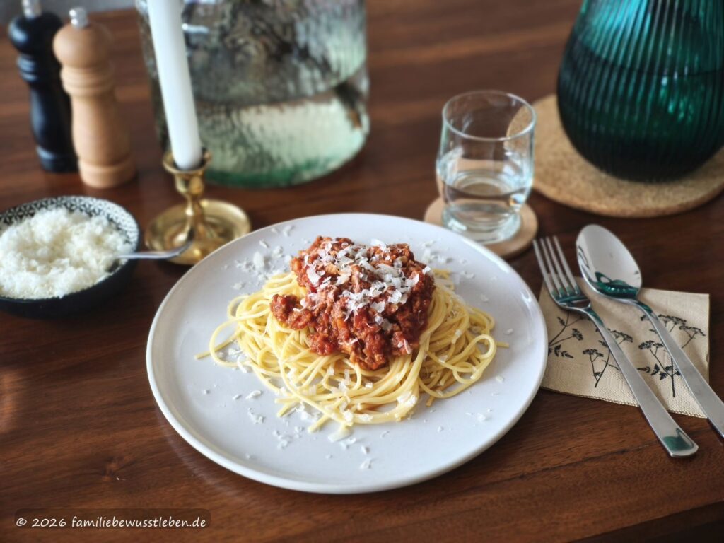 Ein Teller Spaghetti mit Gemüsebolognese, daneben Besteck, ein Glas Wasser, geriebener Käse sowie Salz- und Pfeffermühle. Im Hintergrund stehen eine Vase und ein dunkelgrüner Wasserkrug.