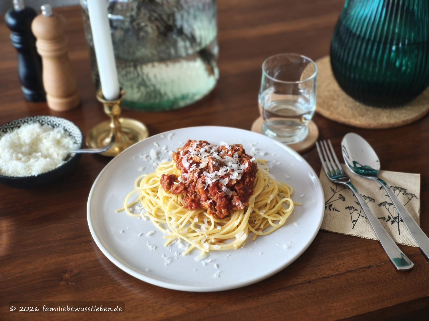 Ein Teller Spaghetti mit Gemüsebolognese, daneben Besteck, ein Glas Wasser, geriebener Käse sowie Salz- und Pfeffermühle. Im Hintergrund stehen eine Vase und ein dunkelgrüner Wasserkrug.