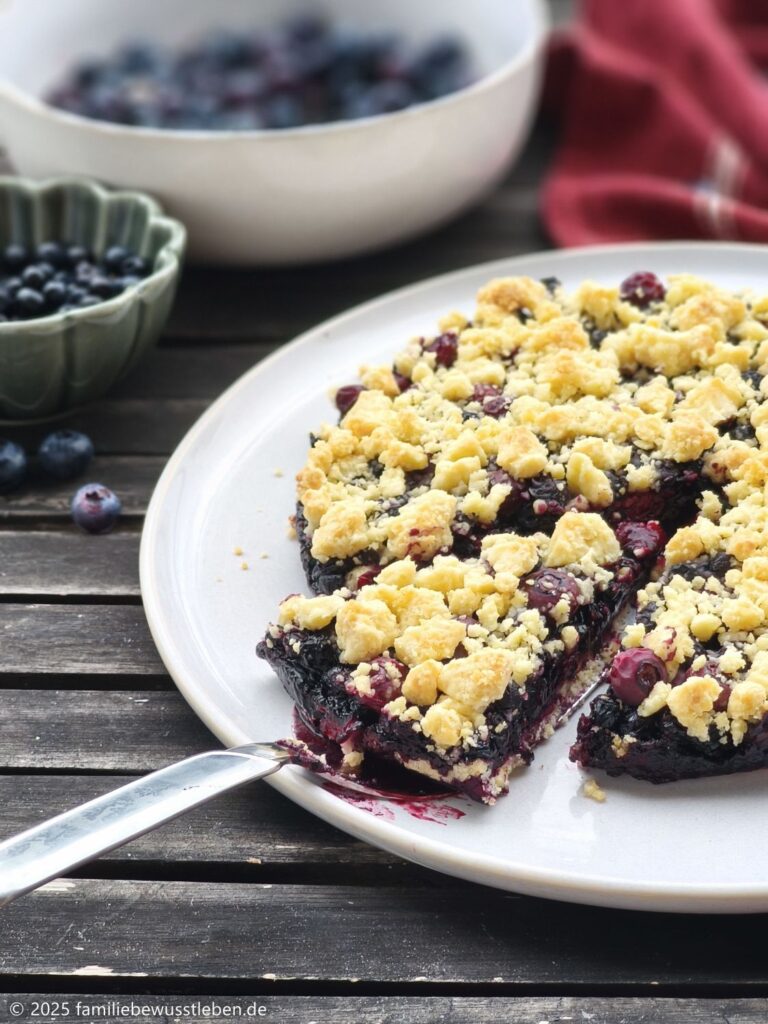 Ein runder Blaubeerkuchen mit Streuseln und ein Stück davon auf einem Kuchenheber. Im Hintergrund Schalen mit Blaubeeren und Waldheidelbeeren.
