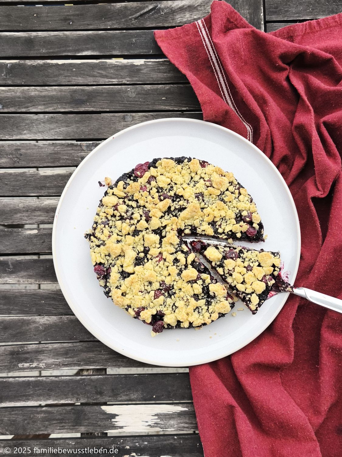 Draufsicht auf Blaubeerkuchen mit Streuseln. Ein Stück ist bereits angeschnitten und wird mit einem Kuchenheber genommen. Ein dunkelrotes Küchenhandtuch liegt neben dem hellen Kuchenteller.