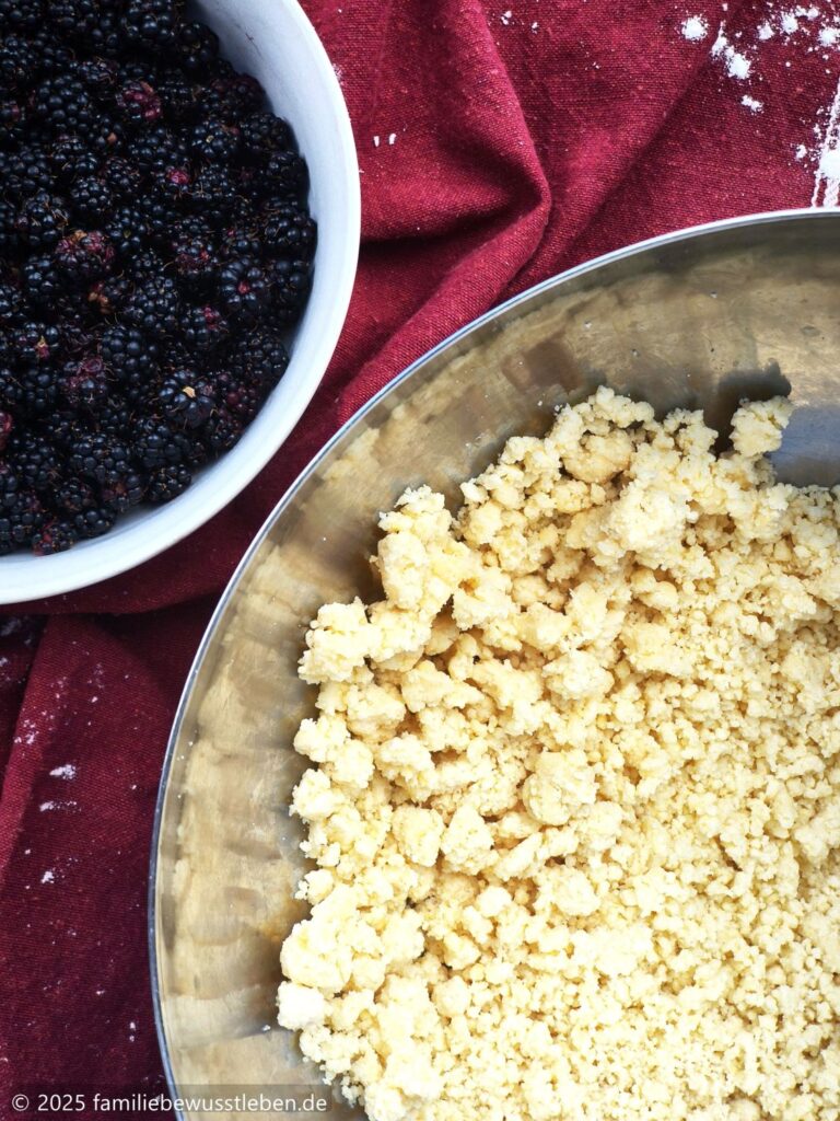 Nahaufnahme von den Streuseln vom Quickteig in einer Edelstahlschüssel und einer Schale mit Brombeeren.