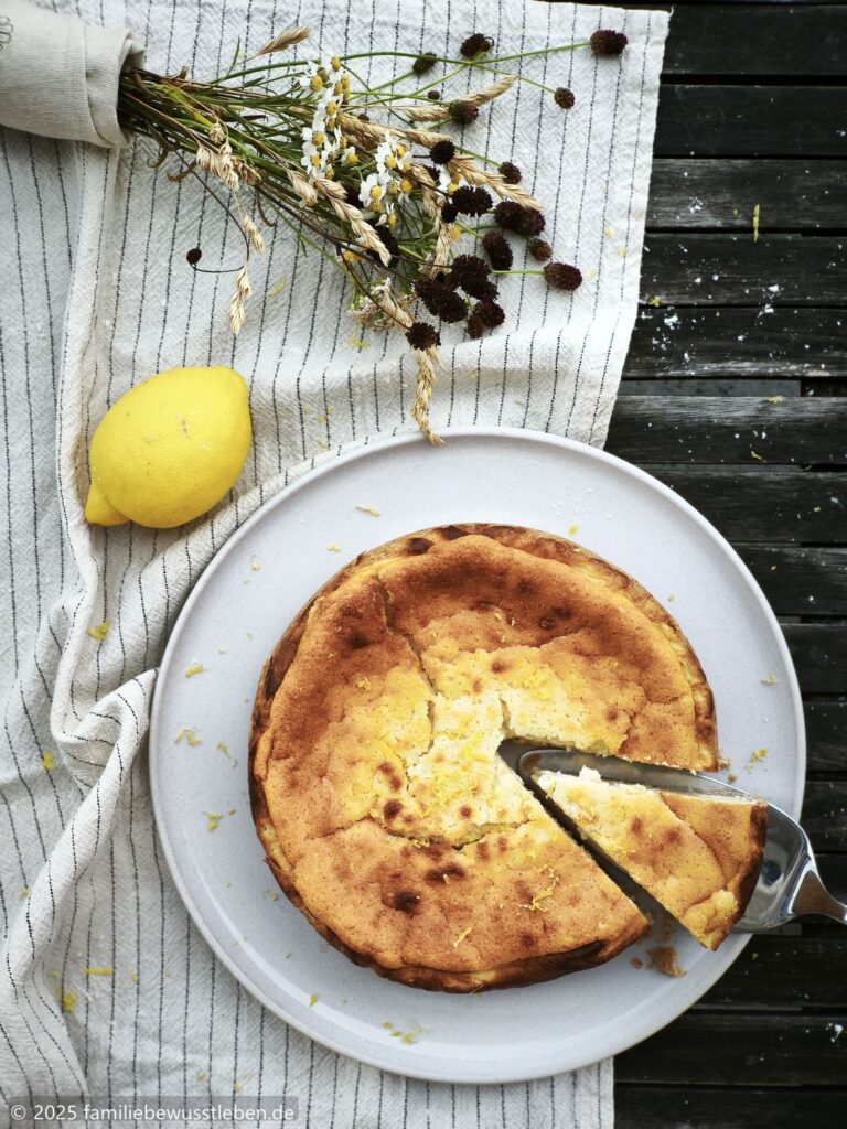 Draufsicht auf einen gesunden Käsekuchen, eine Zitrone und einen Trockenblumenstrauß. Der runde Käsekuchen ist frisch angeschnitten.