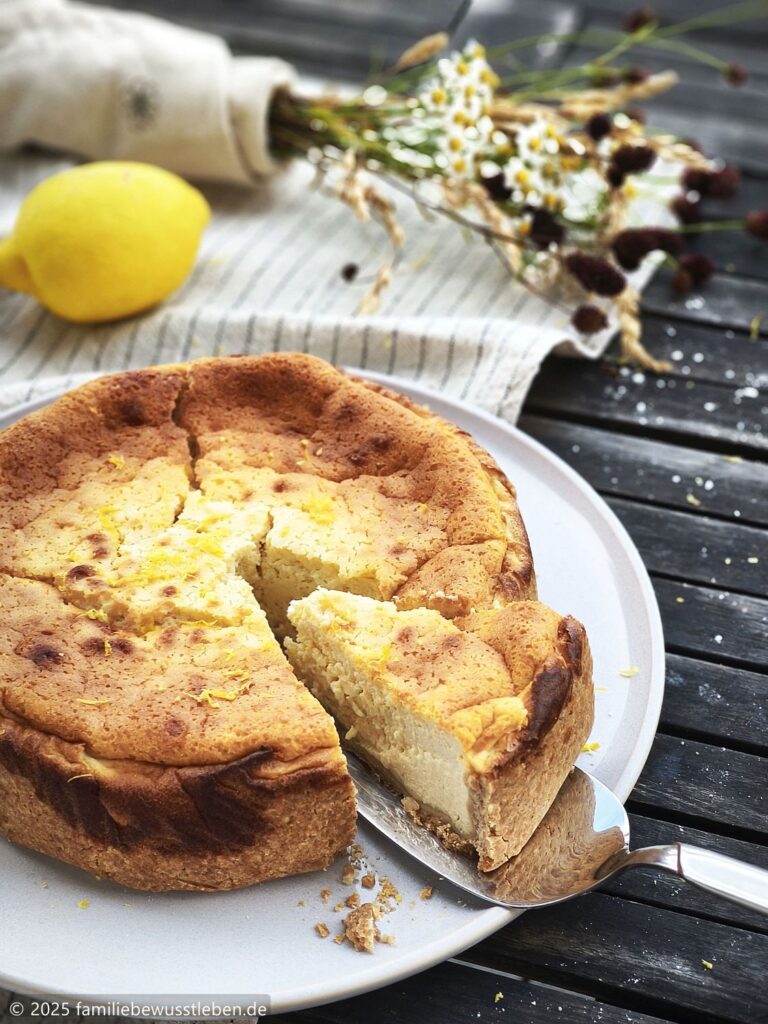 Ein frisch angeschnittener runder Käsekuchen im Vordergrund. Eine Zitronen und sommerlicher Trockenblumen im Hintergrund.