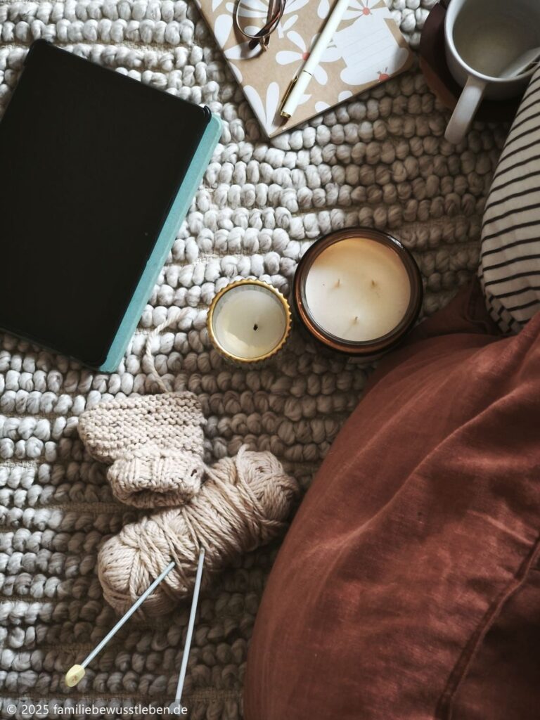 Ein hochkant aufgenommenes Bild zeigt ein Tablet, ein Notizbuch, eine Duftkerze, eine Tasse Tee sowie ein Wollknäuel mit Stricknadeln und einen angefangenen gestrickten Babyschuh. Umgeben von gemütlichen Kissen entsteht eine entspannte Atmosphäre für einen Filmabend in der Schwangerschaft.