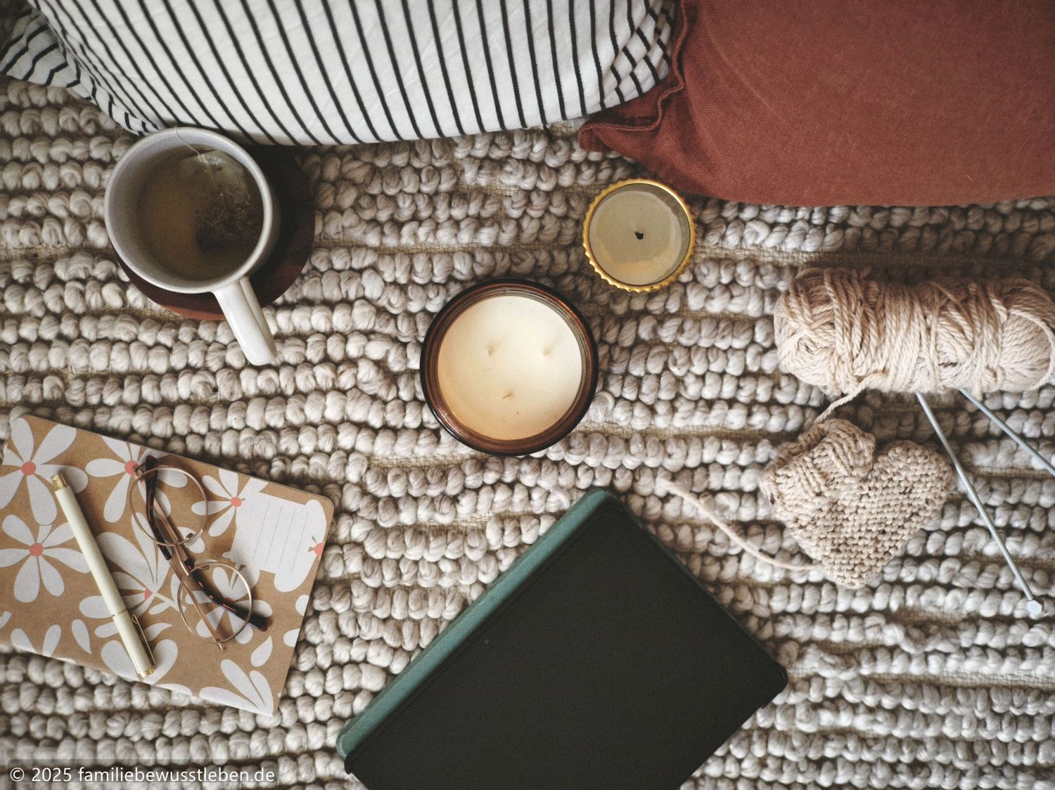 Ein Tablet, eine Duftkerze, eine Tasse Tee, ein Wollknäuel mit Stricknadeln und ein angefangener gestrickter Babyschuh liegen zwischen gemütlichen Kissen – eine entspannte Atmosphäre für einen Filmabend in der Schwangerschaft.